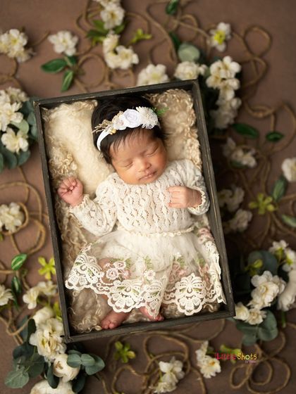 A beautiful full-length shot of a baby in a vintage-style lace dress, sleeping in a rustic box surrounded by white flowers.