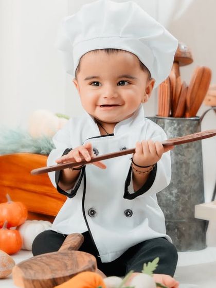 Meet the little chef. This adorable baby is ready to cook up some fun, dressed in a chef's uniform and surrounded by a kitchen full of props.