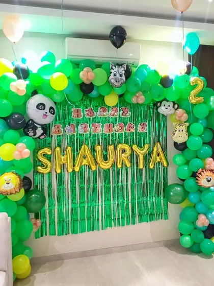 A lush jungle-themed birthday decoration for a two-year-old. I created a full balloon arch in shades of green to mimic a jungle canopy, accented with cute animal foil balloons like a panda, zebra, and lion. The green shimmer backdrop and gold foil name "SHAURYA" make this a fun and adventurous photo corner.