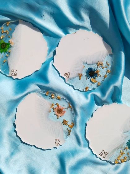 A set of four personalized resin coasters. The design combines a solid white base with a translucent section filled with gold flakes and a variety of colorful preserved flowers, each with a custom initial.