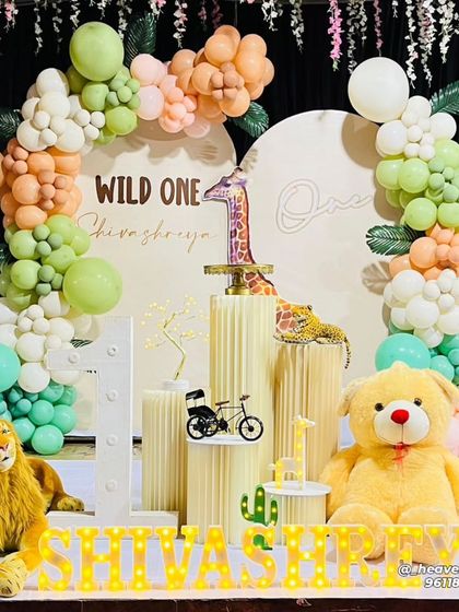 A soft and gentle take on the "Wild One" theme, using pastel green and peach balloons. The setup features a large teddy bear and lion plushie alongside a circular backdrop and fluted plinths for the cake display.