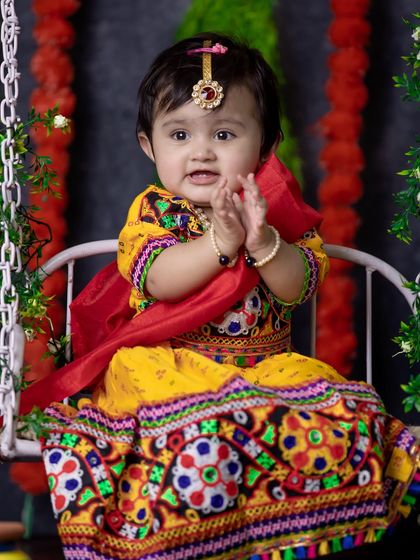 A sweet portrait of a baby girl dressed in a traditional Gujarati-style outfit for her Radha-themed shoot, clapping her hands with joy on a decorated swing.