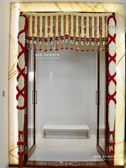 Another view of the white marble pooja room entrance, showcasing the intricate 'X' pattern of the rose garlands on the sides.
