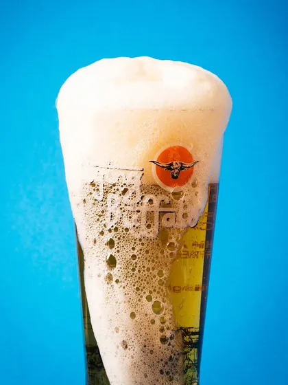 A glass of Flying Buffalo beer with an overflowing head of foam against a bright blue background. It's a fun, energetic shot that screams refreshment.