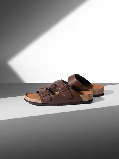 A minimalist shot of brown sandals using strong light and shadow. The graphic composition highlights the product's simple, clean lines and timeless design.