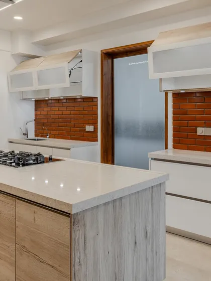 This kitchen in Ahmedabad balances modern geometry with earthy warmth. The textured brick-tile backsplash adds character against the minimal high-gloss lacquer cabinets, while the wood-finish island anchors the space, creating a kitchen that feels both pared-back and expressive.