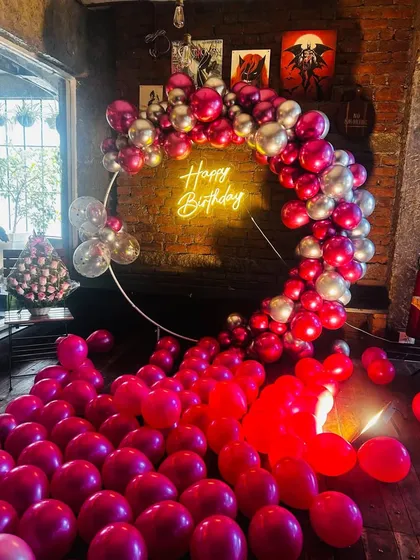 An indoor birthday party setup featuring a large ring backdrop. The circular frame is decorated with a dense arch of red and silver balloons and a central 'Happy Birthday' neon sign, with extra balloons covering the floor for a fun, immersive effect.