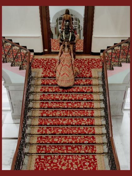 A high-angle shot of the bride and her entourage on the grand red-carpeted staircase. The symmetry and rich colors create a visually stunning and opulent image.