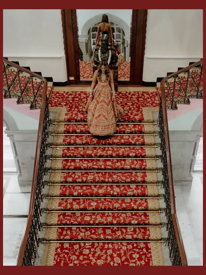 A high-angle shot of the bride and her entourage on the grand red-carpeted staircase. The symmetry and rich colors create a visually stunning and opulent image.