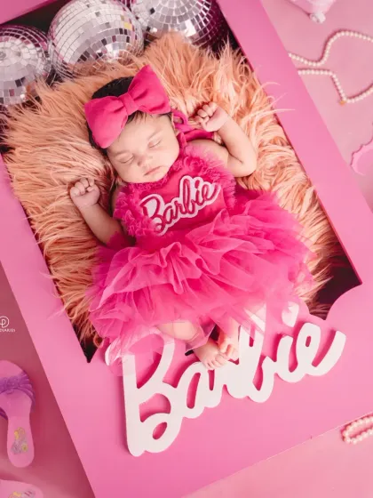A close-up of a sleeping newborn styled for a 'Barbie' themed shoot. She's dressed in a frilly pink outfit inside a custom Barbie box, showcasing the detailed props I use for themed newborn photography.