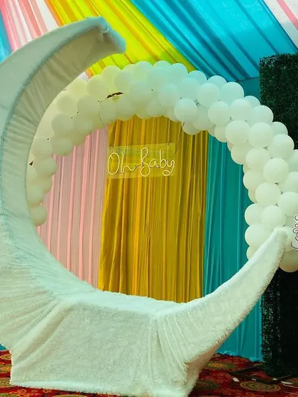 A close-up of a white moon-shaped seating prop for a baby shower, with a Little Krishna cutout and a white balloon arch.