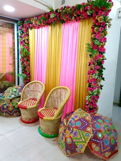 A simple engagement or Haldi corner decorated with a pink and yellow drape, a floral frame, and traditional mooda chairs.
