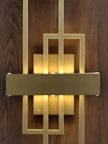 A close-up view of the fine details that elevate our designs. This image showcases a custom-designed geometric wall sconce with a brushed gold finish, mounted on a rich wood veneer panel, adding a touch of artistic flair.