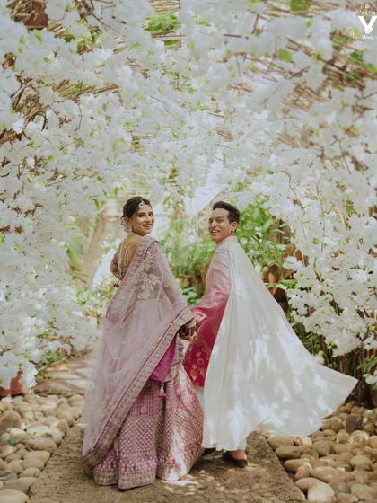 The couple walks through a stunning archway of white flowers, looking back with a smile. The groom's cape adds a touch of modern royalty to this beautiful, fairy-tale-like scene.