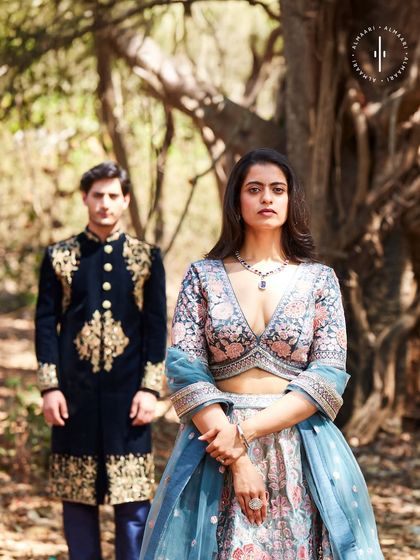 A perfect couple's look. The floral lehenga with a blue dupatta complements the groom's deep blue velvet sherwani with gold embroidery. Renting coordinated outfits is a great way to make a statement together at any wedding.