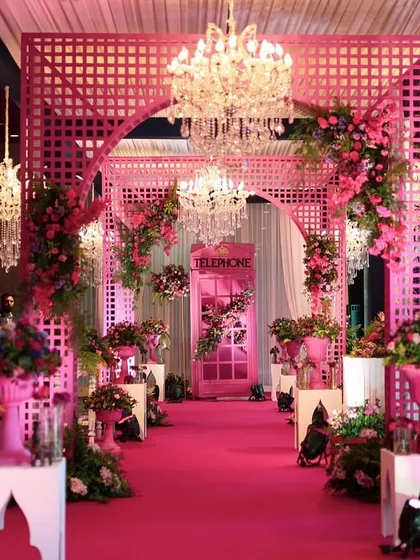 A stunning entrance for a pink-themed Haldi or Mehendi, with a floral-covered archway, crystal chandeliers, and a pink carpet.