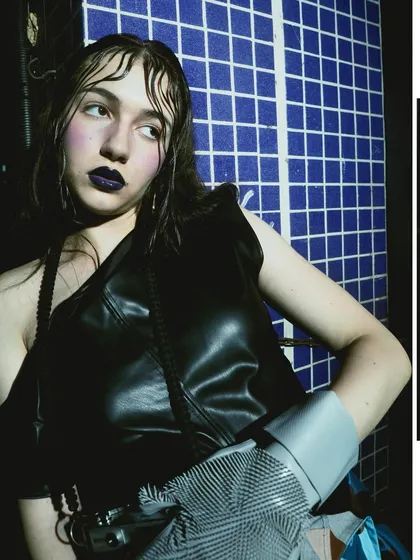 A high-contrast shot against a blue tiled wall, using hard light to create deep shadows. The model's dark makeup and leather top contribute to the edgy, urban aesthetic.