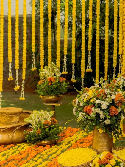 A detail shot of our minimalist Haldi decor. A curtain of marigold strings creates a beautiful photo backdrop, while arrangements of white and orange flowers in brass pots add a touch of elegance.