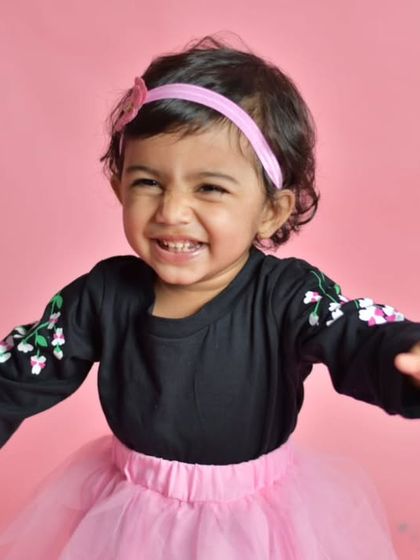 A young girl in a pink tutu and black top beams with a wide, happy smile, her arms open as if ready for a hug, radiating pure happiness.