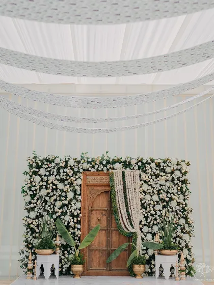 A beautiful and minimalist backdrop for a Haldi ceremony. A wall of white flowers provides a clean and elegant setting, with a traditional wooden door adding a touch of rustic charm.