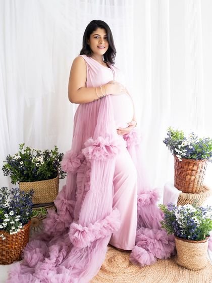 A beautiful solo shot of a mom-to-be in a ruffled pink gown, surrounded by baskets of flowers in a bright, airy studio.