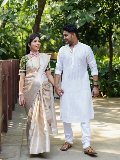 An expecting couple walks hand-in-hand during their outdoor baby shower photoshoot. They are dressed in elegant traditional Indian attire, with the mom-to-be in a beautiful off-white saree.
