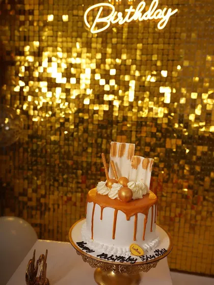 A close-up of the birthday cake, perfectly complementing the decor. The cake sits on a gold stand in front of the shimmering backdrop and neon sign, showing how all the elements work together.