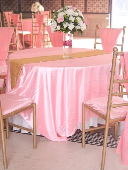 A close-up of the guest table setting, featuring pink satin tablecloths, a gold sequin table runner, and a lovely floral centerpiece. The chairs are adorned with matching pink drapes.