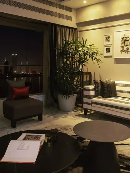 An evening view of the living room, showcasing how the space transforms at night. The lighting design creates a warm, inviting ambiance, with the city lights of the Queen's Necklace providing a spectacular backdrop.