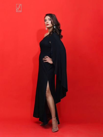 A powerful and elegant studio shot against a bold red background. The model wears a black one-shoulder gown with a dramatic sleeve, creating a striking silhouette.