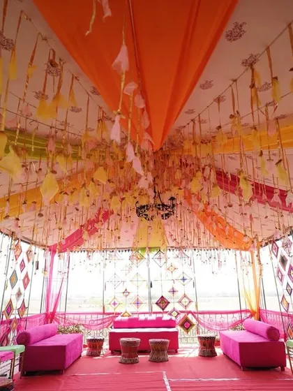 A vibrant and traditional Mehendi setup. The tent is decorated with colorful drapes in shades of orange, yellow, and pink, with intricate hangings that add a festive and cultural touch to the space.
