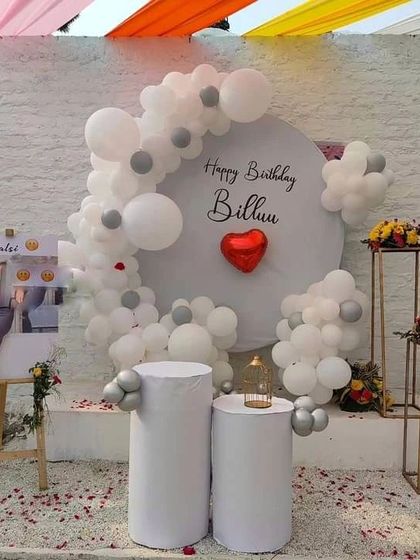 A simple and elegant outdoor birthday setup. This decoration features a personalized circular backdrop, a white and silver balloon garland, and floral arrangements on pedestals.