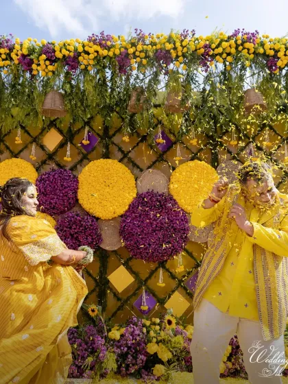 A playful moment from the Haldi ceremony. The bride and groom engage in a fun flower fight in front of a stunning floral backdrop of yellow and purple.