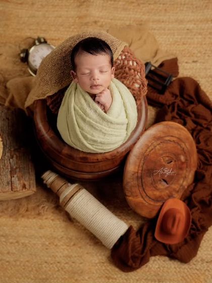 An overhead shot of a newborn sleeping in a wooden bowl, surrounded by rustic props like logs and textured brown fabrics.