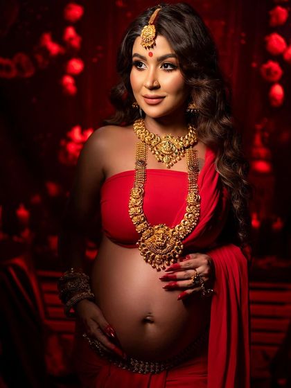 A close-up portrait highlighting the intricate gold jewelry and the serene expression of the mother-to-be. The rich red background and soft lighting enhance the regal and divine feel of the shoot.