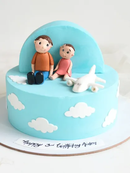 A sweet travel-themed cake featuring handmade fondant figures of a father and child sitting among the clouds with an airplane. This cake has two delicious tiers of milk chocolate and black forest.