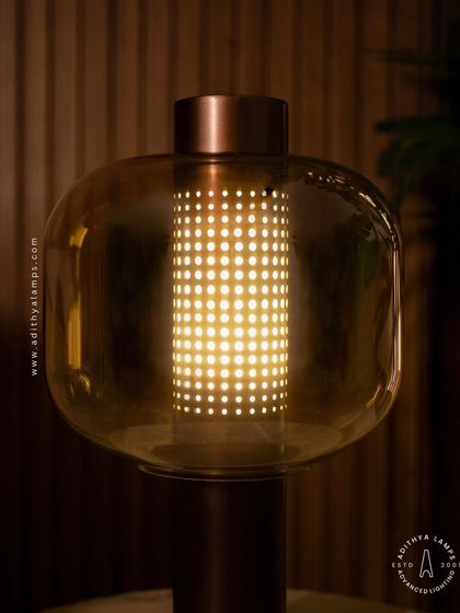 This table lamp features a perforated inner cylinder shielded by an outer amber glass shade. The design creates a mesmerizing pattern of light, offering a warm and intricate glow for a side table or console.