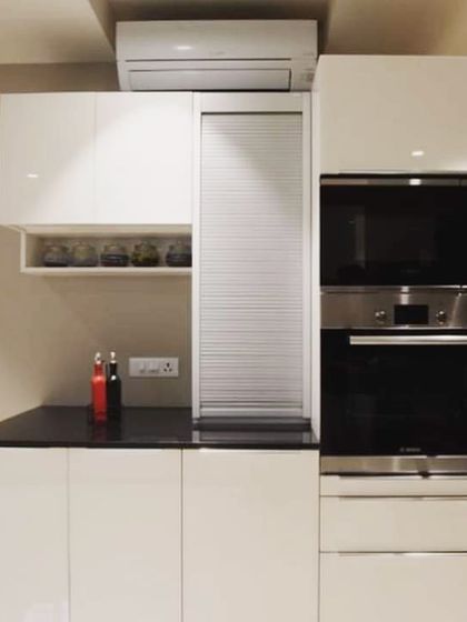 This compact kitchen layout in Lajpat Nagar features a tall unit with a rolling shutter to neatly conceal appliances. The all-white, high-gloss finish makes the space feel larger and brighter.