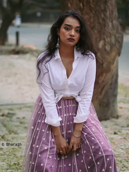 A seated portrait that highlights the elegant flow of the embroidered lavender skirt. The urban park setting gives the outfit a contemporary, wearable feel.