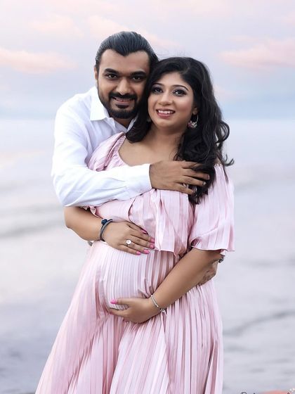A classic couple pose during a serene beach photoshoot. He holds her from behind as they both smile at the camera, standing in the gentle waves with a beautiful sunset sky.