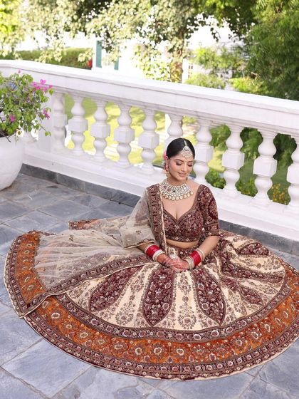 The bride seated, showcasing the beautiful flare and detailed border of the beige and maroon lehenga. The outdoor setting highlights the richness of the colors.