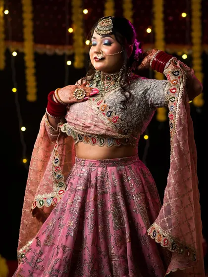 A beautiful bridal look with a soft pink lehenga and dramatic purple-toned eye makeup, set against a festive bokeh background.