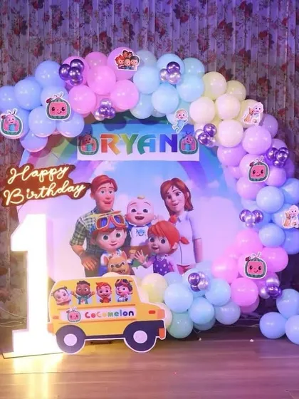 This Cocomelon first birthday party features a pastel balloon arch, character standees, and a large marquee number '1'. The backdrop has all the main characters from the show.