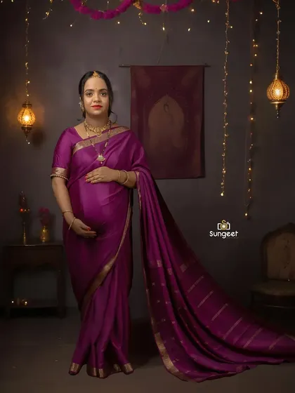 A full-length portrait of a mom-to-be in a rich purple saree. The simple, elegant setup with hanging lanterns puts the focus entirely on her radiant glow.