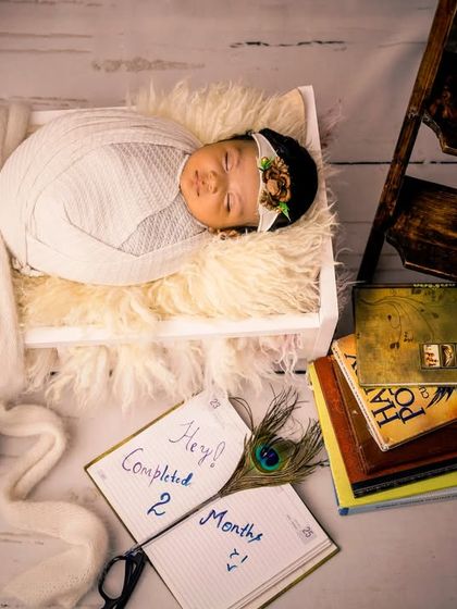 This newborn is sleeping in a tiny white crate, surrounded by books and letters. The setup tells a story, marking the baby's two-month milestone in a creative way.
