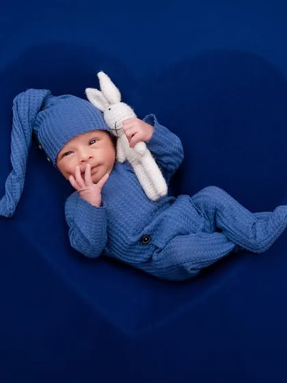 An awake newborn in a blue outfit holds a crochet bunny, looking thoughtful inside a heart-shaped prop.