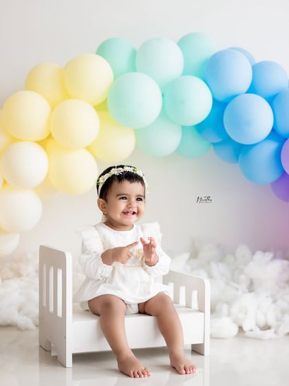 A smiling baby girl sitting on a miniature bed in front of a pastel rainbow balloon arch.