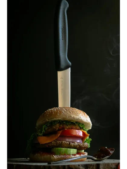 A gourmet chicken burger presented with a knife, set against a dark, smoky background. This dramatic styling gives the burger a bold and premium feel for a restaurant menu.