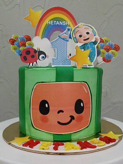 A square Cocomelon cake designed to look like the character's face, with a rainbow and balloons on top.