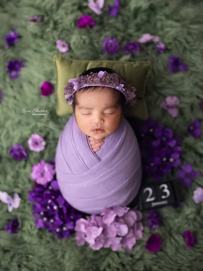 A perfectly wrapped newborn lies on a pillow, surrounded by purple hydrangeas, with a block showing the date of the shoot.